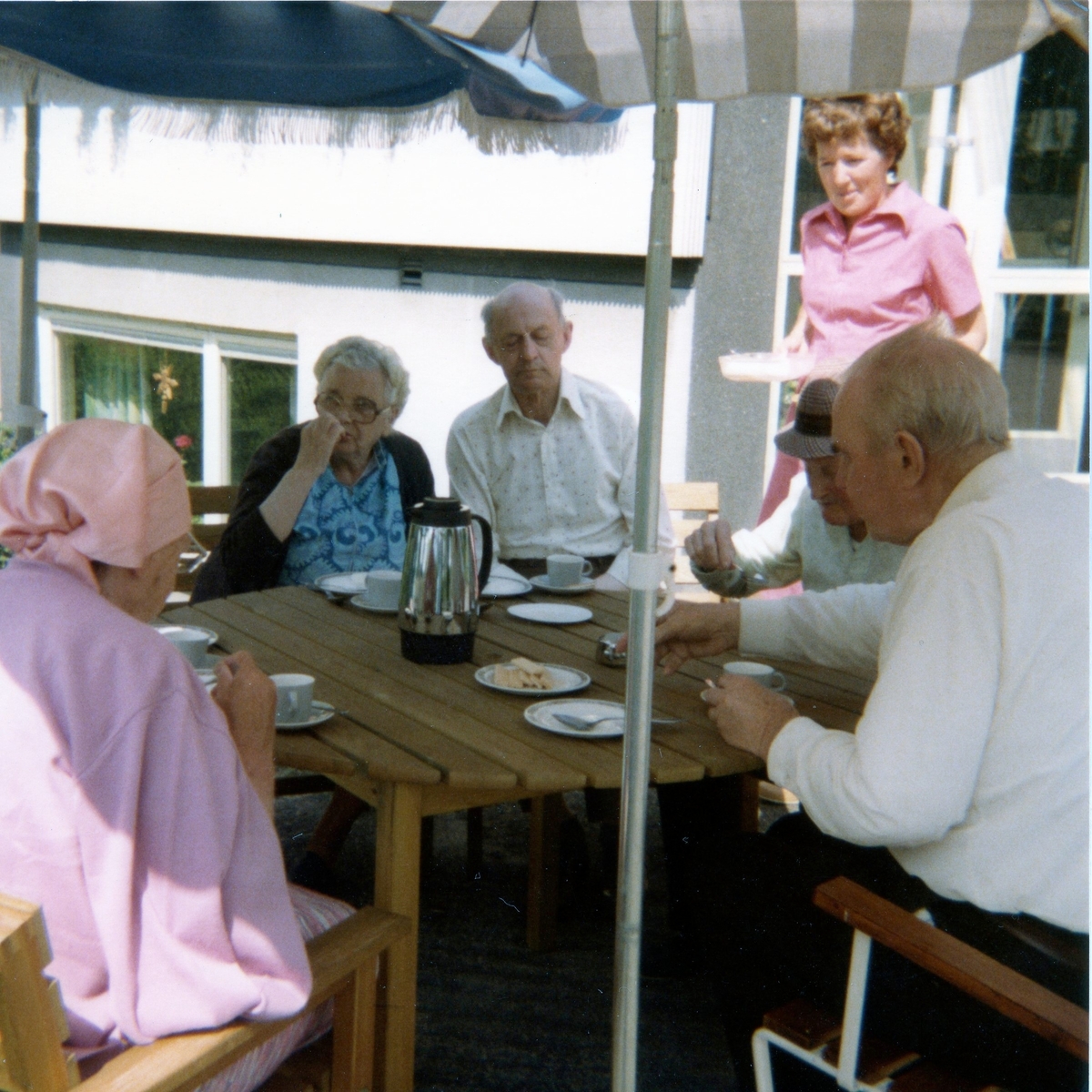 Midsommar på Brattåshemmet, vilken avnjuts utomhus med kaffe och tårta år 1980. Tredje person från vänster är Anselm Zackrisson, kokerska Ingela Sjöberg samt Alfred Mattsson (iklädd hatt). Övriga är okända.