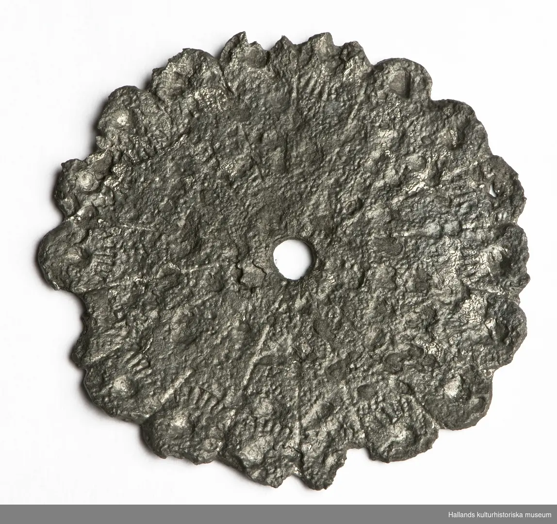 En cirkelformad platta med 18 stycken runda taggar i kanten. I metallen syns rester av mönster och i mitten finns ett hål. Troligtvis fårn likkista.
