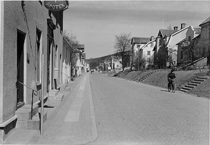 Brahegatan norrut från hörnet Postgränd, närmast i bild fastighet med Apotek. En äldre man med hatt kommer cyklande söderut. Trottoaren har en rad med borgmästarsten.