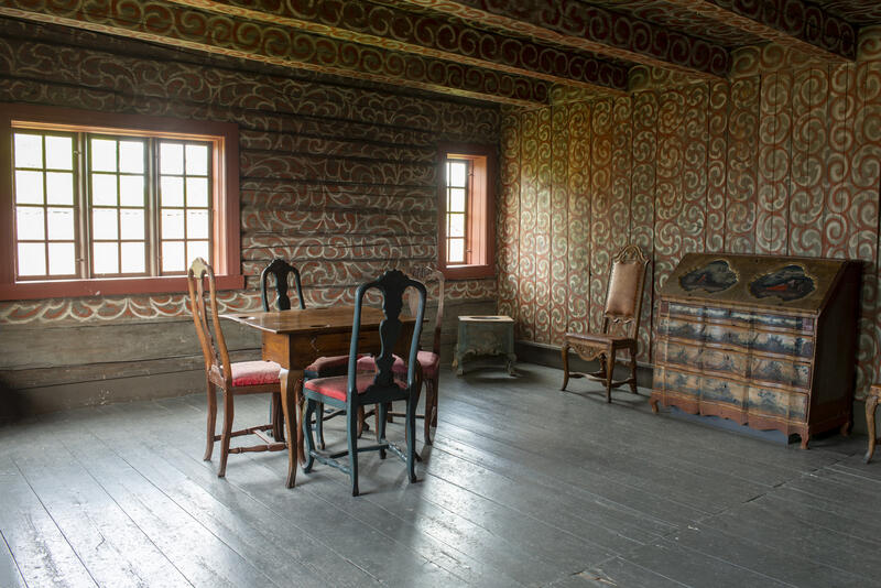 Interiør fra Bolstadbygningen viser en stolgruppe og en kiste i et rom med dekormalte tømmervegger.