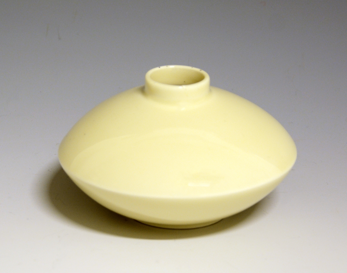 Liten vase i porselen. Liten fotrand og lav sylindrisk hals. Elfenbensfarget glasur. Nora Gulbrandsens modell 2129, tegnet i 1935.