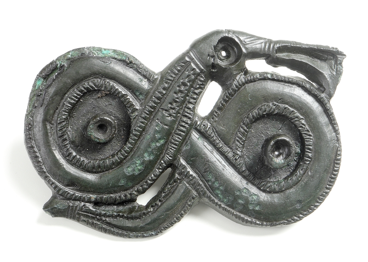 Dräktfibula i form av ett ormöglespänne av Cu-legering. Ormfigur som slingrar likt en åtta. I de två öglorna, samt i det nedsänkta "ögat" har det antagligen suttit stenar. På baksidan finns rester av fästanordningen.