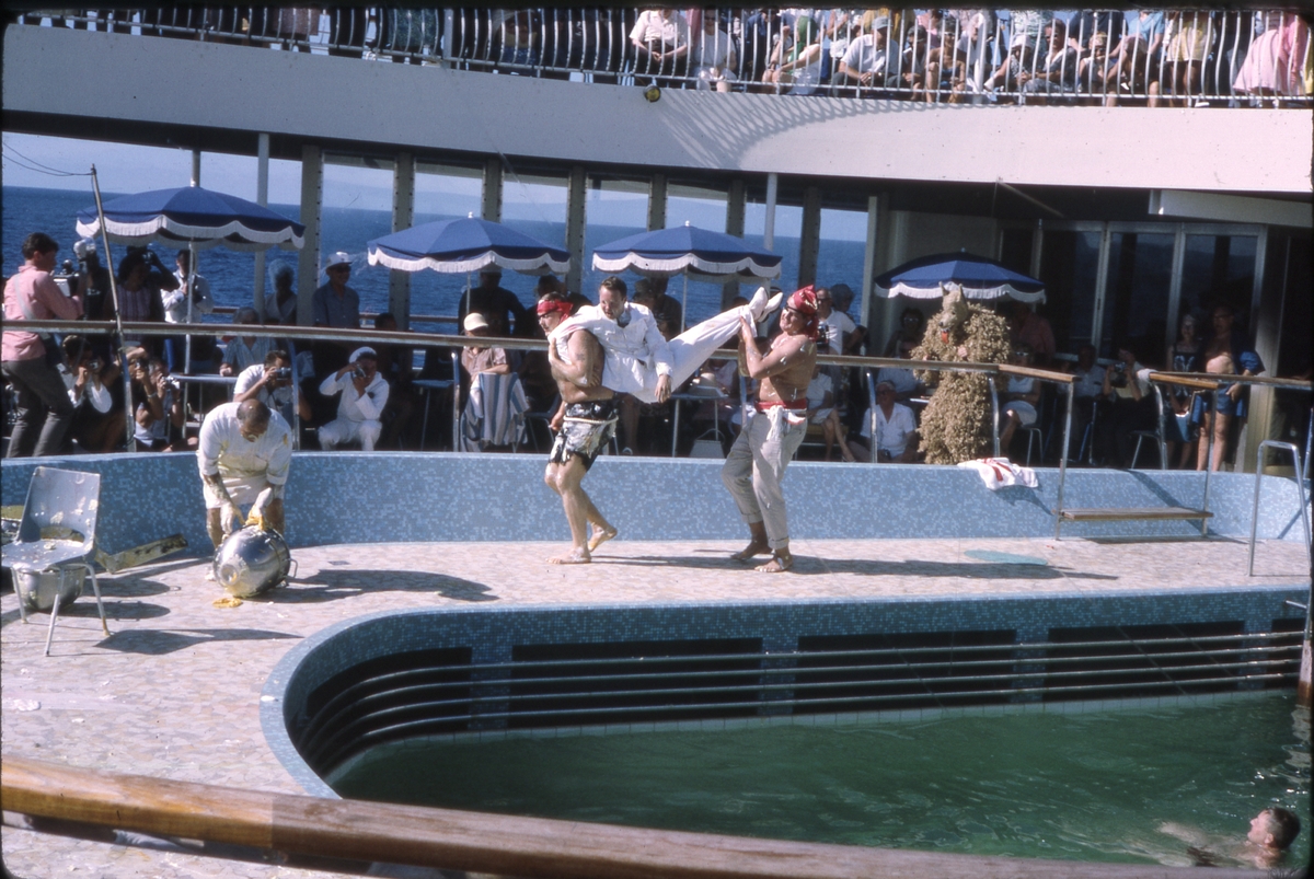 Ekvatordåp om bord på 'M/S Sagafjord'. To menn utkledd som pirater bærer dresskledd mann langs bassengkant. Skuelystne, fotografer og mann utkledd som ulv i bakgrunnen. 'Sagafjord' Around The World via Africa Cruise 1966.