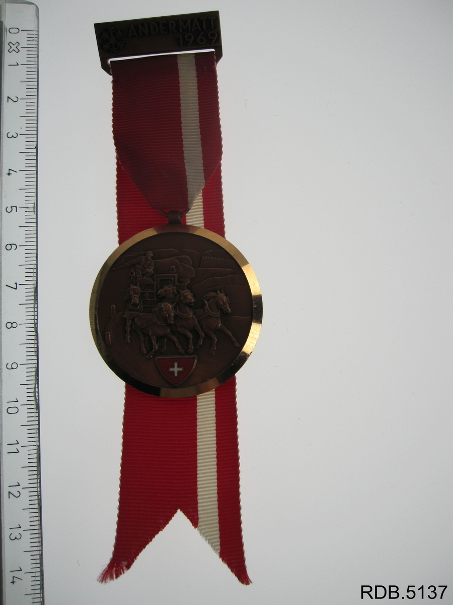 Rund medalje festet til et bånd. Ei firkantet plate er festet i andre enden av båndet.  Innskrift på baksiden av medaljen, motiv på forsiden. Innskrift på platen, ei sikkerhetsnål på baksiden av platen.
