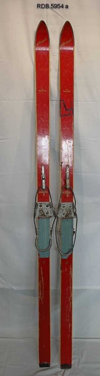 Et par røde, laminerte barne-treski med stålkanter og stålbeslag bak. På venstre ski er det noe malingssøl. Kandaharbinding og blå fotplate.