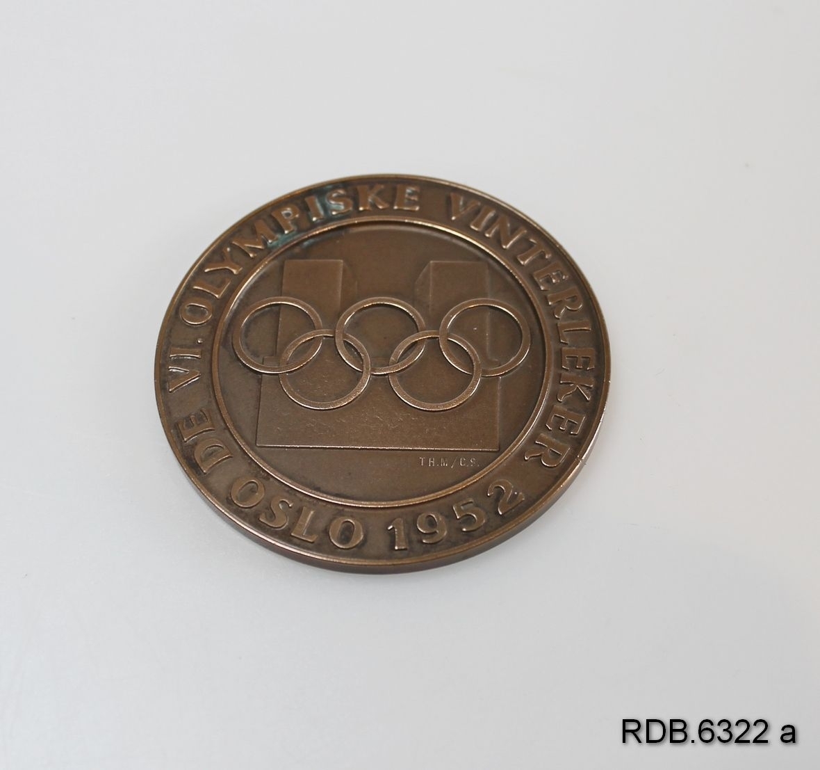 Deltakermedalje fra OL i Oslo 1952. Medaljen er rund med de olympiske ringer og påskrift på framsida. På baksida: En stor snøkrystall i midten, 3 små snøkrystaller rundt kanten pluss påskrift.