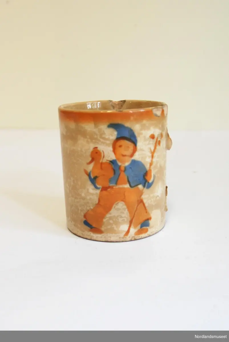 Kopp i porselen med motiv av gutt som holder en and og en kjepp. Mangler håndtak og har flere skader. Misfarget. Under står det: "Utenlandsk u1851 21".