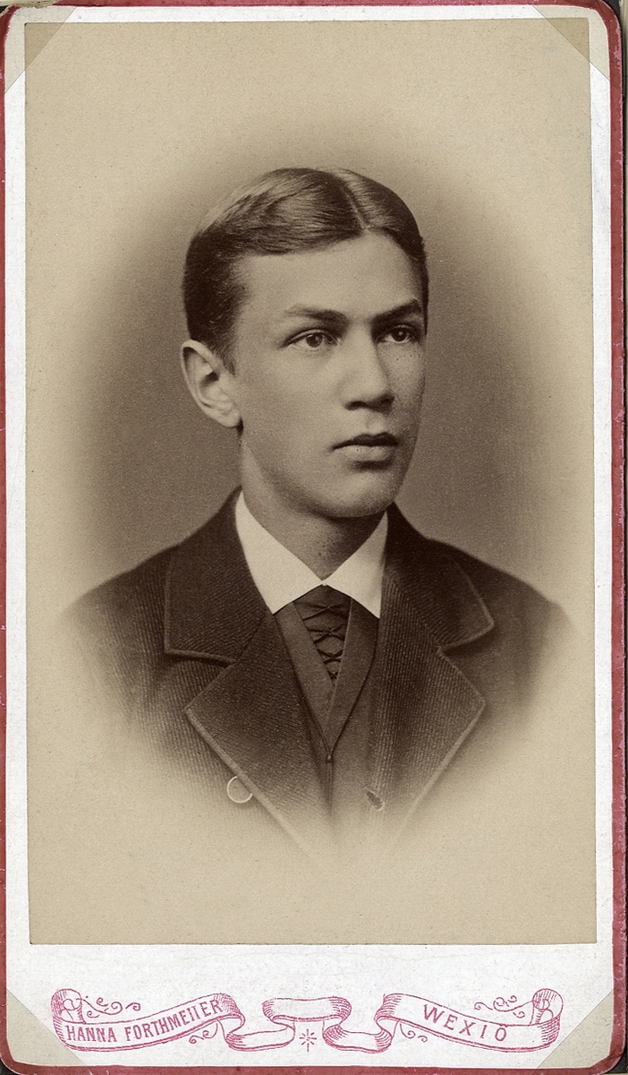 Porträttfoto av en ung man i mörk kavajkostym med väst m.m. 
Bröstbild, halvprofil. Ateljéfoto.