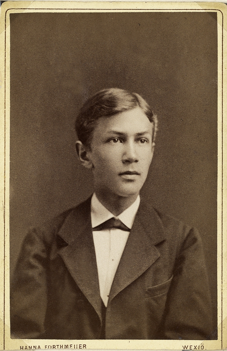 Porträttfoto av en ung man i mörk kavajkostym med väst m.m. 
Bröstbild, halvprofil. Ateljéfoto.