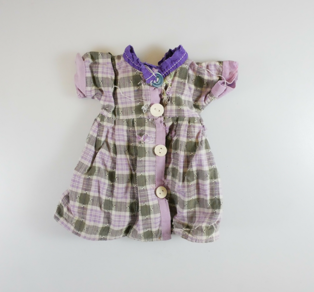 Kortermet liten kjole i lilla og grønnrutete mønster, antageligvis brukt som dukkeklær. Tre hvite og en blå knapp på forstykker. 
