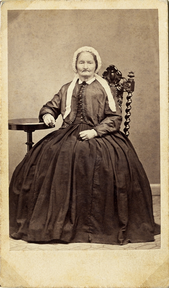 Porträttfoto av en medelålders kvinna i krinolin med jacka och spetsmössa med långa band på huvudet. Hon sitter på en högryggad stol vid ett pelarbord.
Helfigur,en face. Ateljéfoto.