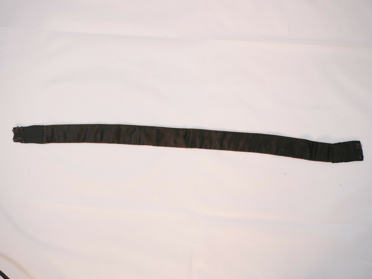 Band av silkesateng, fora på baksida med bomullstoff (svart med kvite prikkar). Skøytt med svart silke i begge endane. To sett krokar og maljer.
