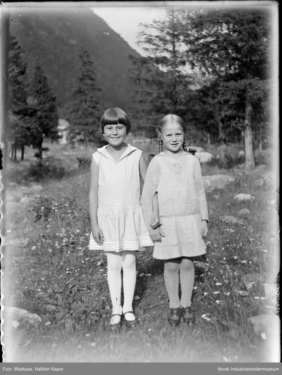 To jenter i kjole stående arm i arm på uteområde.