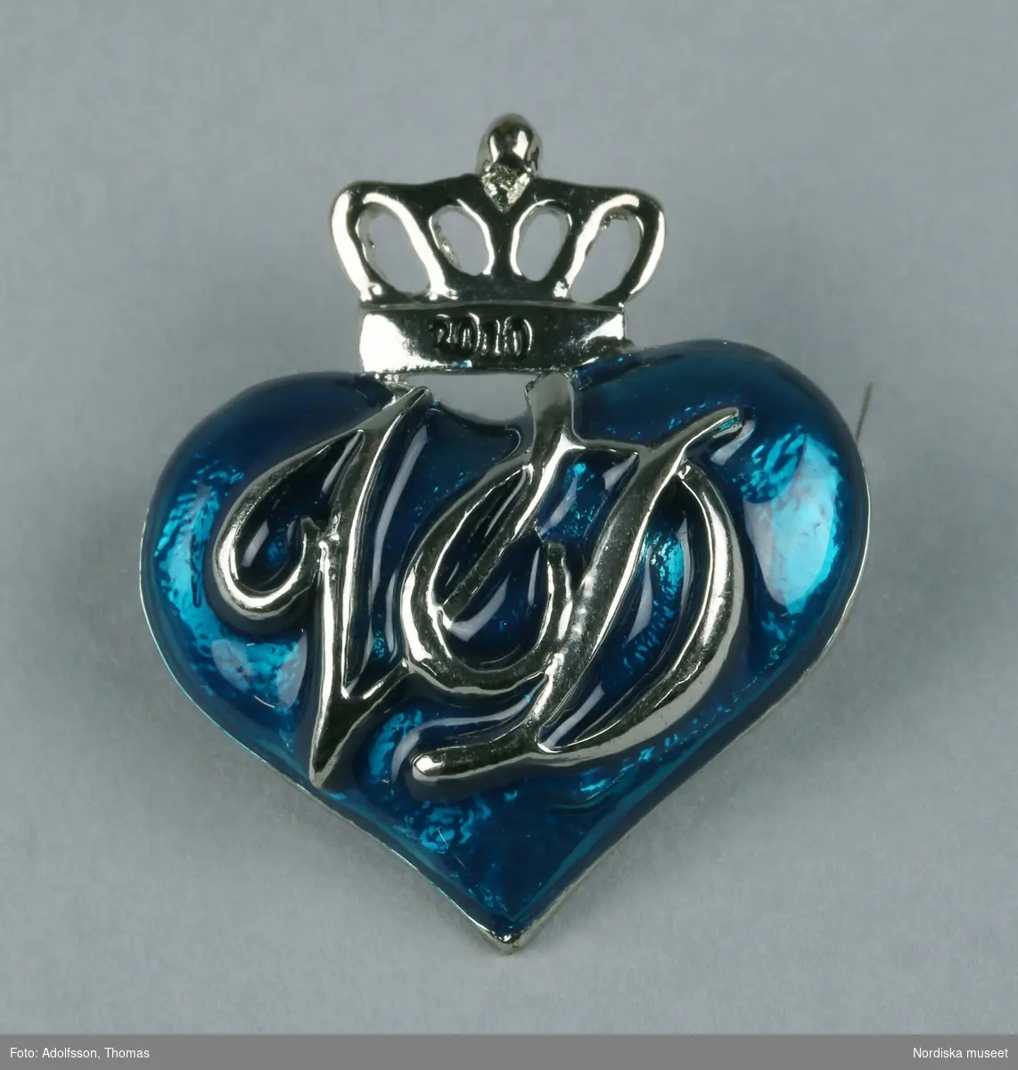 Brosch av vitmetall i form av blått hjärta med sluten krona ovantill. På kronan text "2010" coh på hjärtat "VD", vilket står för Viktoria och Daniel. På baksidan nål. 
/Leif Wallin 2010-09-10