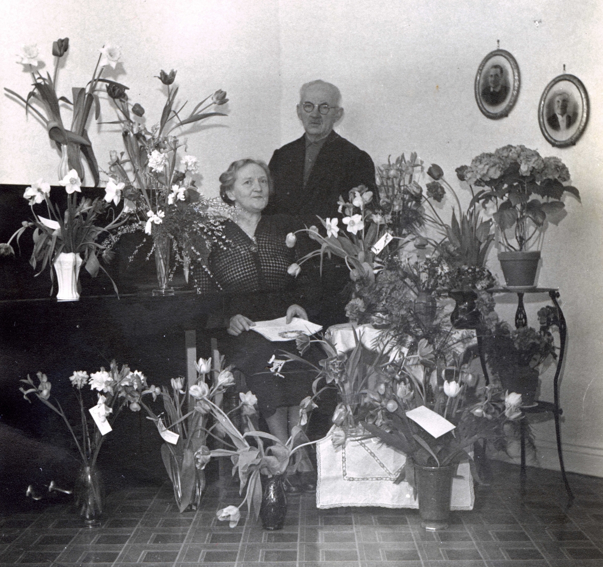 Judit Johansson fyller 60 år och uppvaktas med blomster i Vommedal Östergård "Olas" år 1953. Brevid står maken Gustav Johansson. Judit föddes 1893 i Bölet och Gustav föddes 1886 i Fässberg. De gifte sig 6/6 1917 och drev jordbruk i Vommedal.