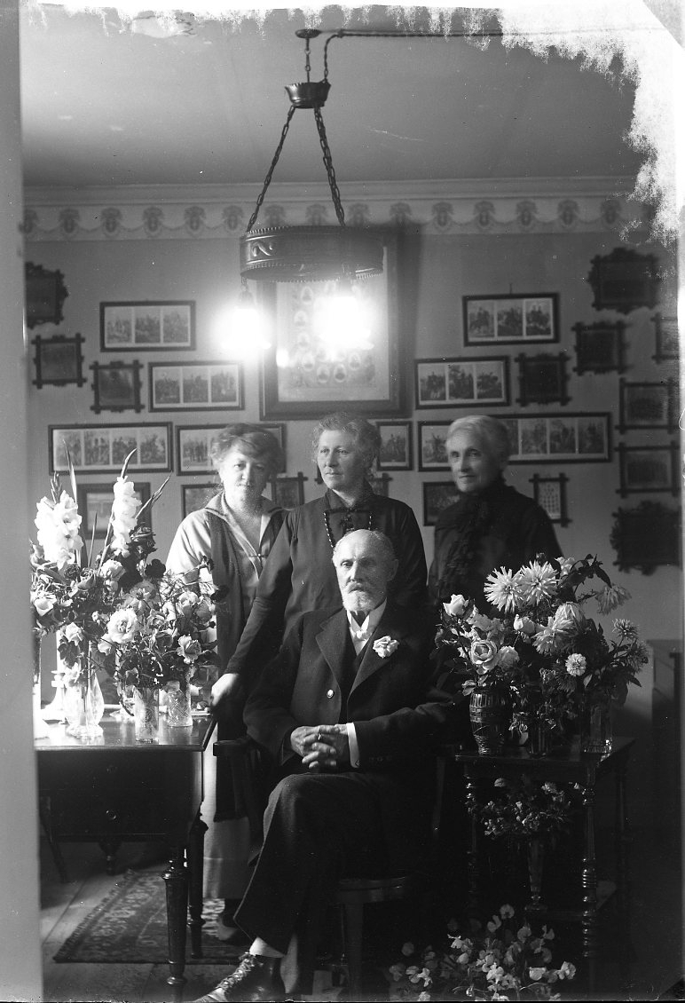 Major Ohlson sitter i en stol i sitt hem omgiven av bord med mängder av blommor. Bakom honom står tre kvinnor. Hon till vänster är Maria von Rosen, född Frick. Hon till höger är sannolikt hans hustru Helena Malvina Frick. Ett stort antal inramade fotografier på väggen bakom honom, troligen militära scener. Fotografi i samband med hans 80-årsdag.