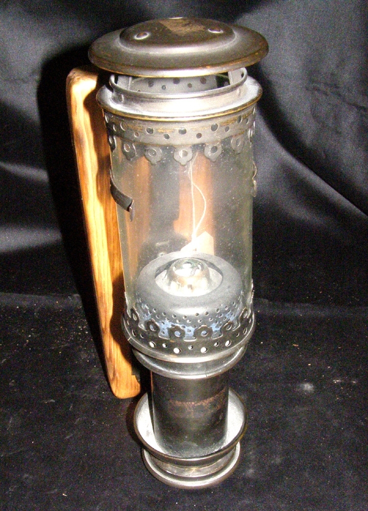 Cylindrisk lampett av metall med rörformad glaskupa. Krön av metall.
Avsedd för stearinljus. En fjäder i hållaren skjuter upp ljuset vartefter det brinner ner.