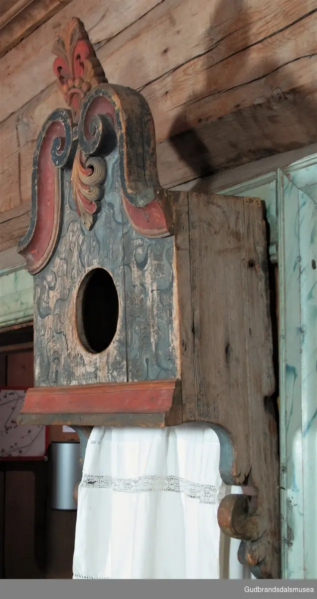 "Utskoren Krone med perleband. Breide Lister måla i rustraudt og blågrønt. Sirkelrund opning" (Katalog for Lom bygdesamling, 1925).
Bostkastet har skåret dekor med et spir sentralt plassert i toppen samt en perlerad i bunnen av spiret.
Skurddekoren kan være av Hans Olson Helleløkken fra Vågå (1791-1858) eller inspirert av ham. Et særtrekk for Helleløkken var skap dekorert med et spir med bladskurd midt på skapet sin topp. Den sentralt plasserte bladskurden på bostkastet representerer samme stiltrekk i mindre format. ”Perleraden” som man kan se i underkanten av spiret har sannsynligvis hentet inspirasjon fra Jakup Rasmusson Sæterdalen (Lom/Vågå, 1756-1821) sin bruk av samme element.