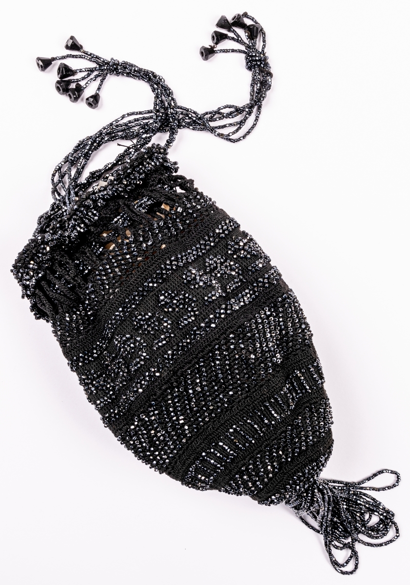 Aftonväska, påsmodell, svart, virkad med inträdda pärlor. Nertill pärlfransar. Väskan bärs i pärlklädda snoddar vilka används för att försluta väskan upptill.