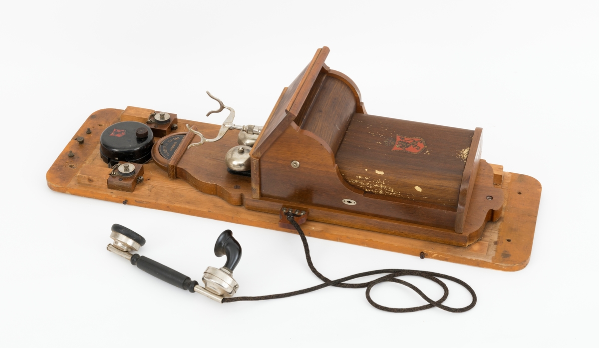 Sveivetelefon, antakelig av type som ble brukt omkring 1. verdenskrig.  Telefonen er montert på ei 1,6 centimeter tjukk treplate.  Apparatet består av ei "kasse" av tre og jernblikk, alt lakkert i edeltreimiterende brunfarge.  I denne delen av apparatet inngår et skrått lokk, en liten skriveklaff, som antakelig skjermer underliggende mekanikk og muligens også batterier.  Ellers har apparatet en forkrommet holder for telefonrøret, som også er forkrommet, men har bakelittkomponenter for håndgrep (midten), øreanlegg og taletrakt.  Apparatet har to omvendt skålformete forkrommete bjeller bak rørholderen, som antakelig gav fra seg klemt når det kom henvendelser per telefon.  Øverst på apparatet er det et slags halvsirkelformet "hode" med informasjon om produsenten.  Her er det også et rødt skjold med en løvefigur med krone over.  Dene logoen finnes også på en svartlakkert, sylindrisk boks på apparatet.  Fra denne boksen er det ledningsforbindelse til stålskiver, som er fastskrudd på begge sider av apparatet. Plata telefonen er montert på har blyantpåskriften «Stor-En-Dammen». Dette antas å være en god indikasjon på at dette apparatet har vært montert i koia ved den nevnte dammen i et av Osensjøens sidevassdrag. Her ble telefonen brukt når fløterne i nedenforliggende deler av vassdraget ønsket å gjøre avtaler som regulerte vanntilførselen i elveløpet på en måte som var optimal ned henblikk på å få tømmeret til å flyte mot Osensjøen.