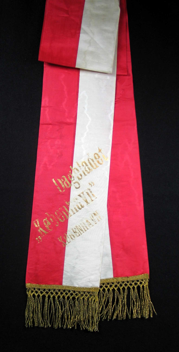 Begravelsessløyfe i hvit og rød silke med frynser og tekst i gull.