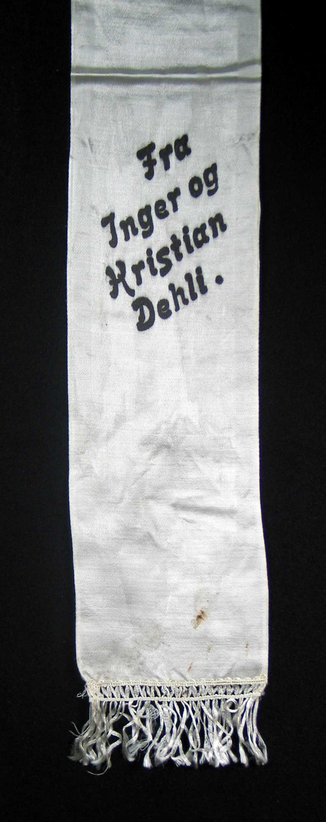 Begravelsessløyfe i hvit silke med svart tekst. Frynser.