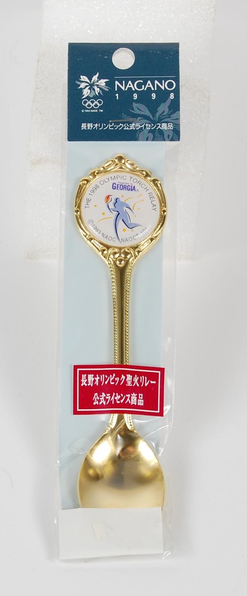 Gullfarget teskje med logo for fakkelstafetten i forbindelse med de olympiske vinterleker i Nagano i 1998. Teskjea er i original innpakning.