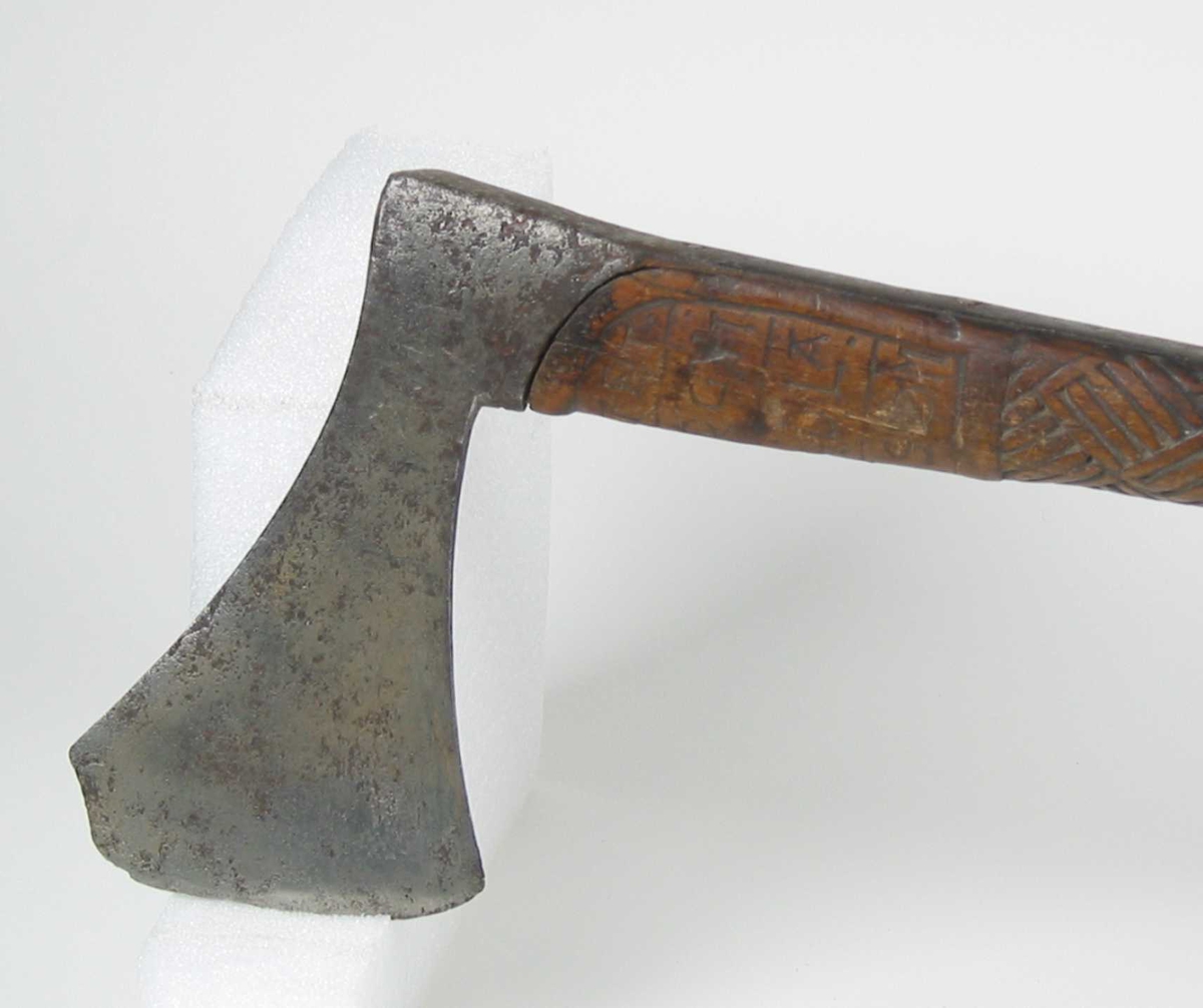 Asymmetrisk økseblad, sammensveiset med overliggende jernskinne.
Fremre øksebladspiss brukket.
Rikt dekorert skaft med flettet rutemotiv. Innskripsjon på øvre del av skaftet (se Påført tekst)
