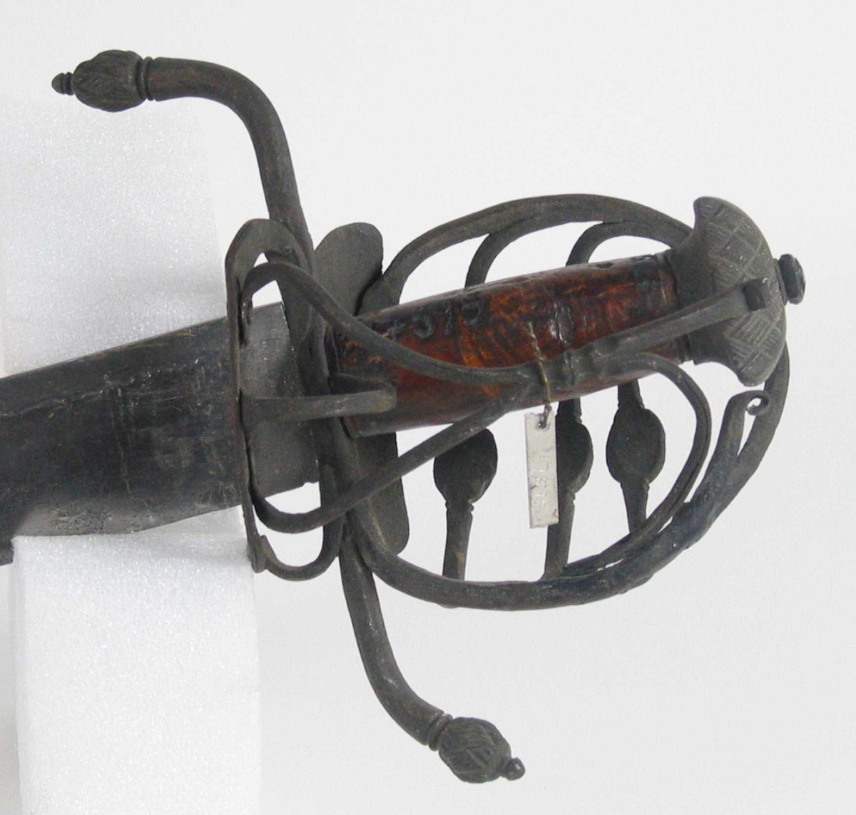 Pyramideformet knapp med kryssfilt dekor og asymmetrisk feste med 3 sidebøyler med fortykninger på midten på utsiden og en på innsiden. Nyreformet parerplate på utsiden og klingebrekker og tommelbøyle på innsiden. S-formede, runde parérstenger med fortykning med filt dekor og dråpeformet avslutning.
Tregrep. Enegget krum, kileformet klinge med en ryggstillt blodrand. Ryggslipt mot spissen. Spiss brukket. Tre hull på spiss. Ett smedstempel.

