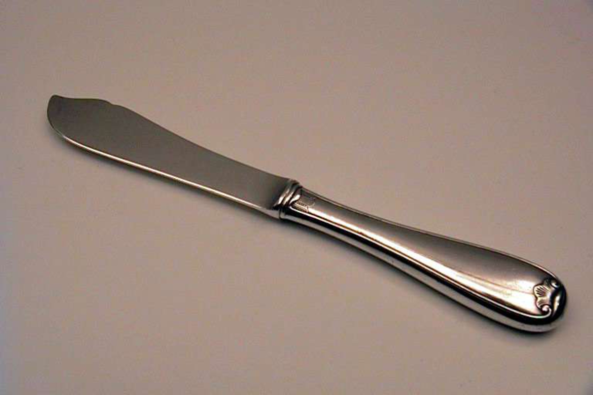 Kniv til fiskebestikk. Materialet er 90 gram sølv. Kniven har et enkelt bladmønster (viftemønster). Stempelet består av et tårn og bokstavene N (i kvadrat) og NM 90.