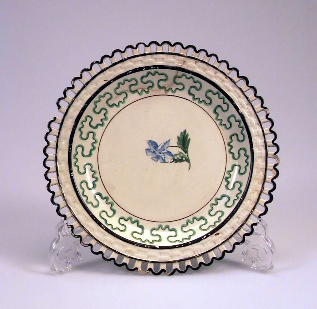 Tallerken i keramikk med kremfarget glasur og med dekor i brunt, grønt og blått. Den har 'hullfallskant'. 