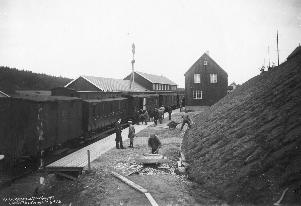 Bygging av jernbanestasjon. Stasjonshus, lokomotivstall, kombinert lastetog og persontog står på stasjonen. Personer på perrongen.