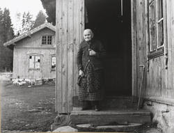 Eldre kvinne i døråpning, spaserstokk,deler av gårdstun