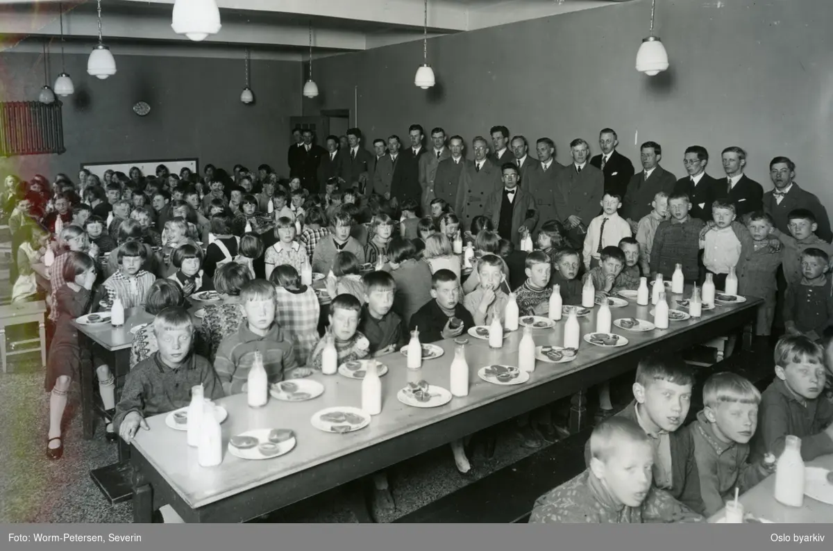 Skolefrokost i matsalen. Elever ved langbord med mat og melkeflasker. Lærere oppstilt langs veggen i bakgrunnen. Forløper til Oslofrokosten (fast etablert 1932). Fra Helserådets arkiv.