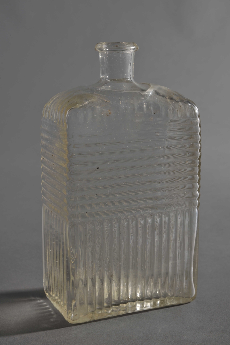 Blank, blåst glassflaske med en antydning av konkav bunn. Rektangulær form, høye avrundete skuldre, kort hals. Vertikale striper på nedre halvdel, og horisontale striper på øvre halvdel, samt striper nedover skuldrene.