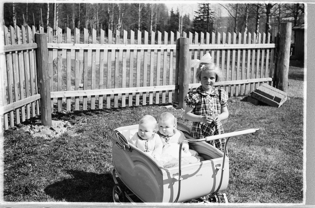 Tvillingene i vogna er Knut og Bjørg Svenskerud f. 1949. Jenta med sløyfe er Inger Svenskerud f. 1945. 
Bildet er tatt ved Åsmarka kirkestue. 
 Jente med sløyfe i håret og to babyer i barnevogn. 