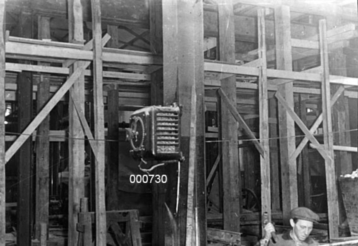 Interiør fra A/S Buskerud Papirfabrikk på Åssida i Drammen (den gang i Lier kommune).  I 1937 ble papirmaskinen ved denne fabrikken fullstendig ombygd, særlig for å kunne kjøre maskinen med større driftshastigheter, slik at en kunne oppnå større produksjonsvolumer.  I den forbindelse la bedriften om fra dampdrift til elektrisk drift.  Fotografiet viser innvendige byggearbeider.  Det er oppsatt kraftige stendere for en ny bindingsverksvegg, og foran den påbegynte veggen står et trestillas.  Helt i forgrunnen ei søyle med et påmontert elektrisk apparat. 