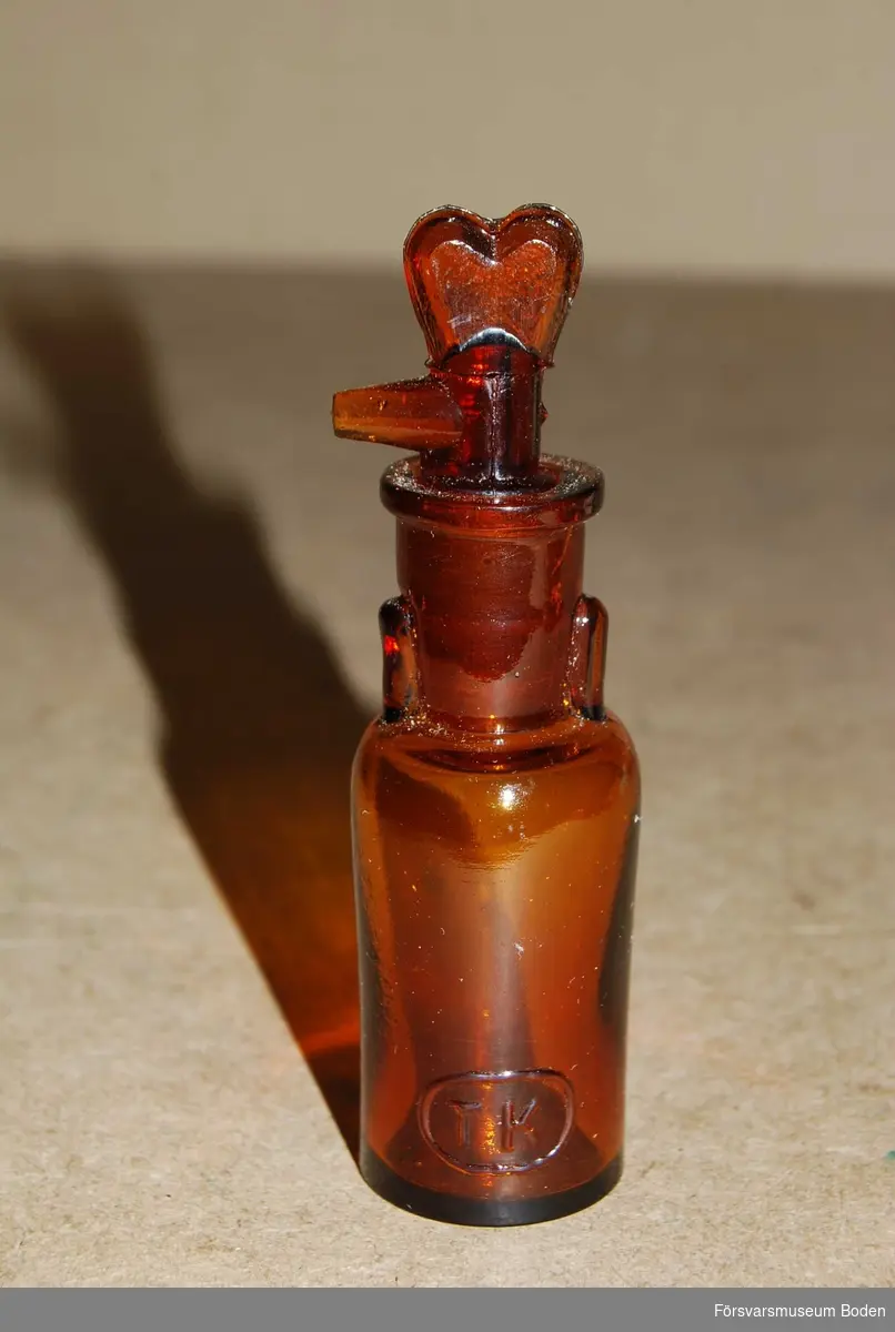 Av brunfärgat glas. Korken av samma material har konisk slipning och en längsgående räffla. Den kan vridas till motsvarande urtag i flaskans hals, varvid en droppfunktion fås.