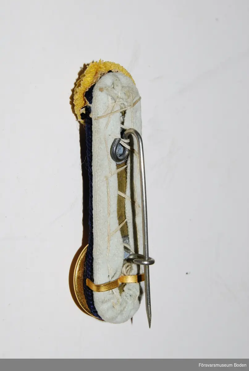 Avlång metallplåt med vitt kläde och fastlödd nål för montering på mössan. Upptill gul sidenkokard och nertill guldfärgad knapp med tre kronor, mellan dessa blå beläggningssnören m/1904.