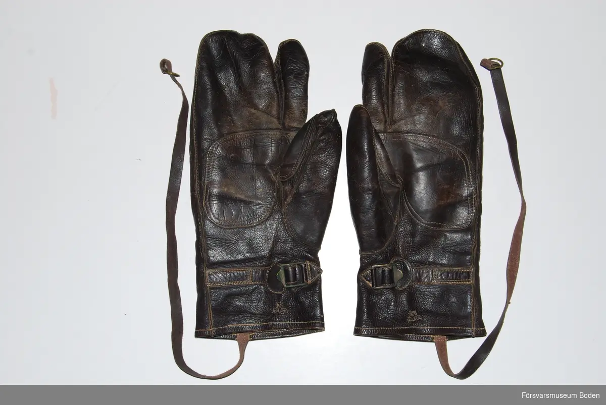 Ett par handskar av trefingersmodell i mörkbrunt skinn. Spännrem runt handleden samt lång rem för fastsättning i fältdräkten. Präglade med tre kronor-symbol. Tillhörande innervantar registrerade under FMB.002605.