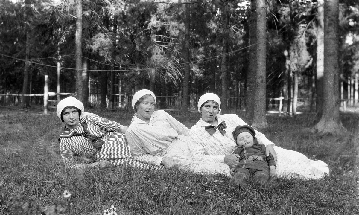 Tre kvinner og et barn som ligger på grasbakken i parklignende omgivelser. Identitet ukjent. Stedet er trolig Gardermoen.