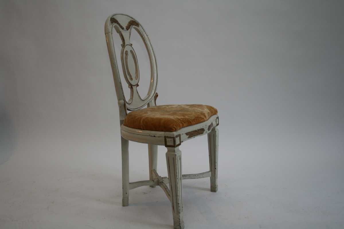 Hvitmalt stol med forgylt dekor. Oval rygg og stoppet sete trukket med gul plysj. Rette bein med kanelleringer, diagonal sprosse. 
Louiz Seize-stil.
