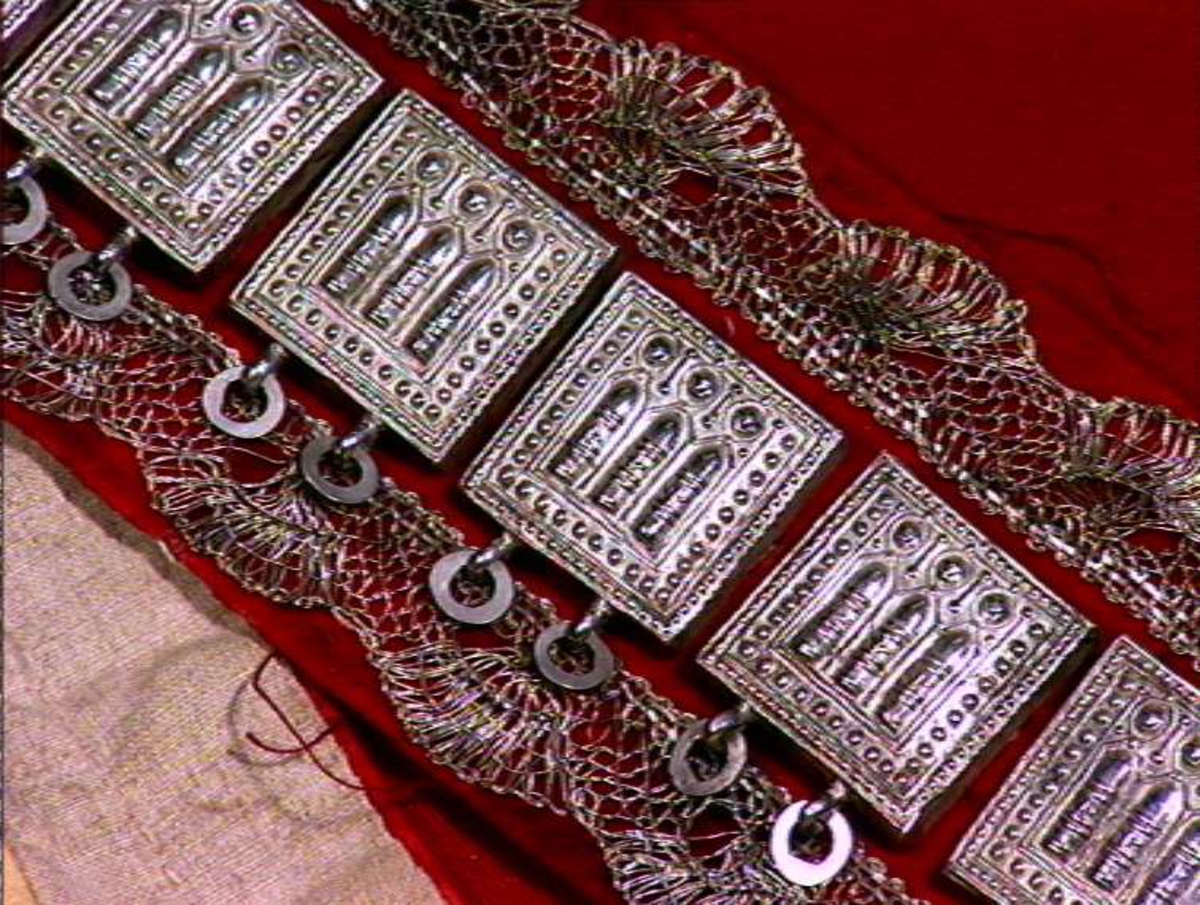 Rødt belte med sprette og sprot.
Stølar av sølv på rødt klede, sølvbeslag og metallknipling.