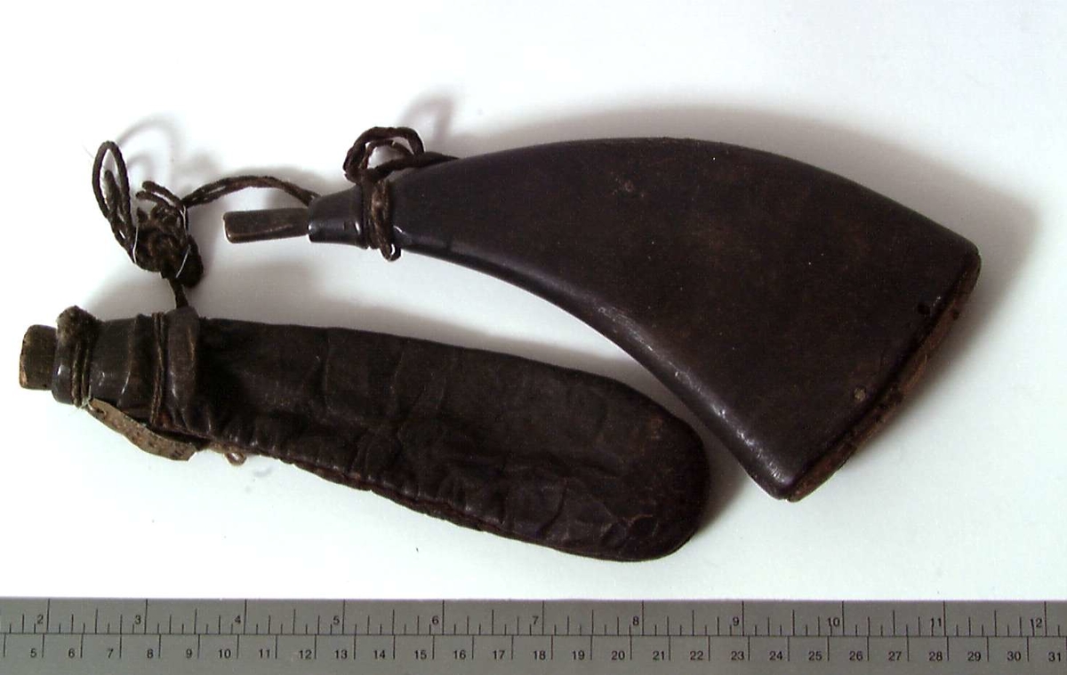 Avflatet, mørk beholder av horn med trepropp, knyttet sammen med en avlang skinnpose med trehals og propp.