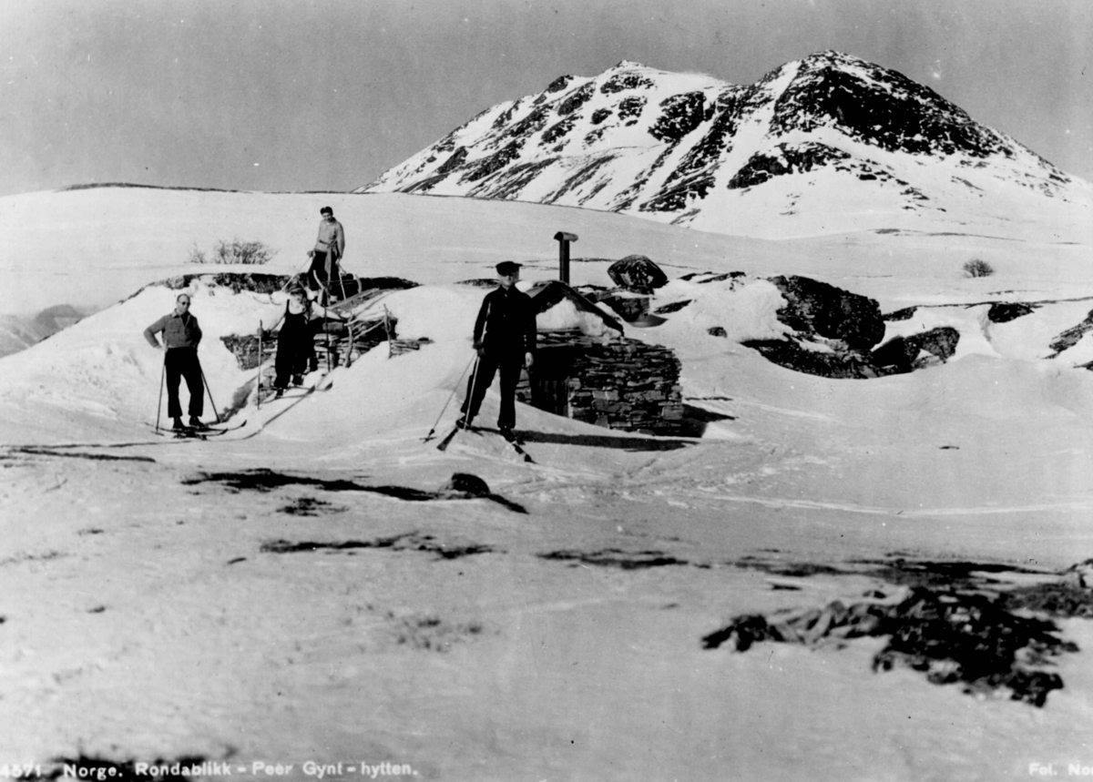 Avfotografert postkort. Tre mannlige og en kvinnelig skiløper foran Peer Gynt-hytta på Rondablikk.