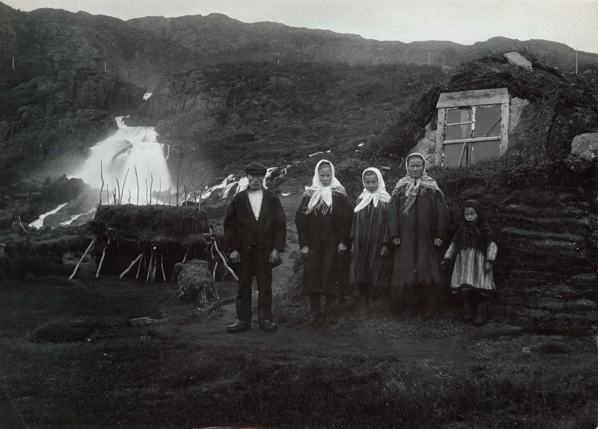 Familien Johansen i samisk drakt foran gamme ved Adamsfjordfossen i Lebesby. Piken til høyre er Valborg.
Del av serie fra en forskningsreise i Øst-Finnmark 1909.