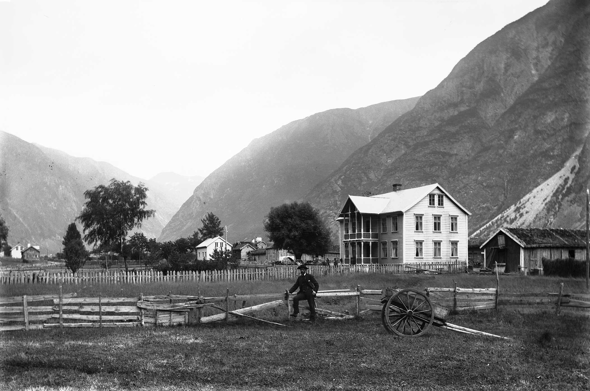 Blaaflat hotell og kyststasjon i fjell- og dallandskap. Mann lener seg mot gjerde, foran står en kjerre.