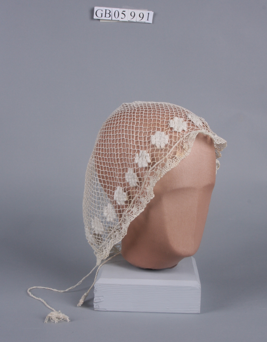 Trukket sammen i kronen. Broderte blomster langs kanten. Kant av tettere masker. Trekkebånd med dusker.