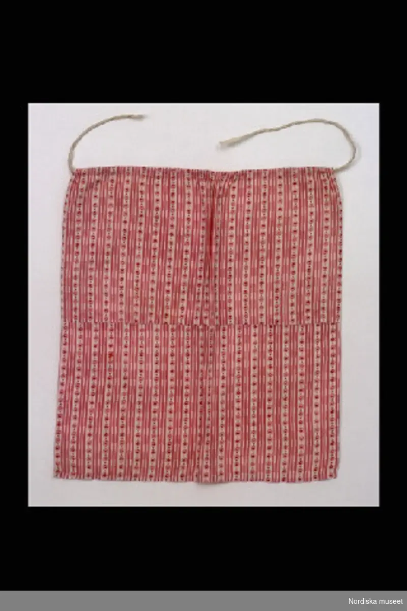 Inventering Sesam 1996-1999:
L  27  cm
Förkläde till docka av småmönstrad rosa kattun. Upptill dragsko, vita knytband av bomull.
Tillhör docka Preciosa inv 111.388.
/Birgitta Martinius 1997