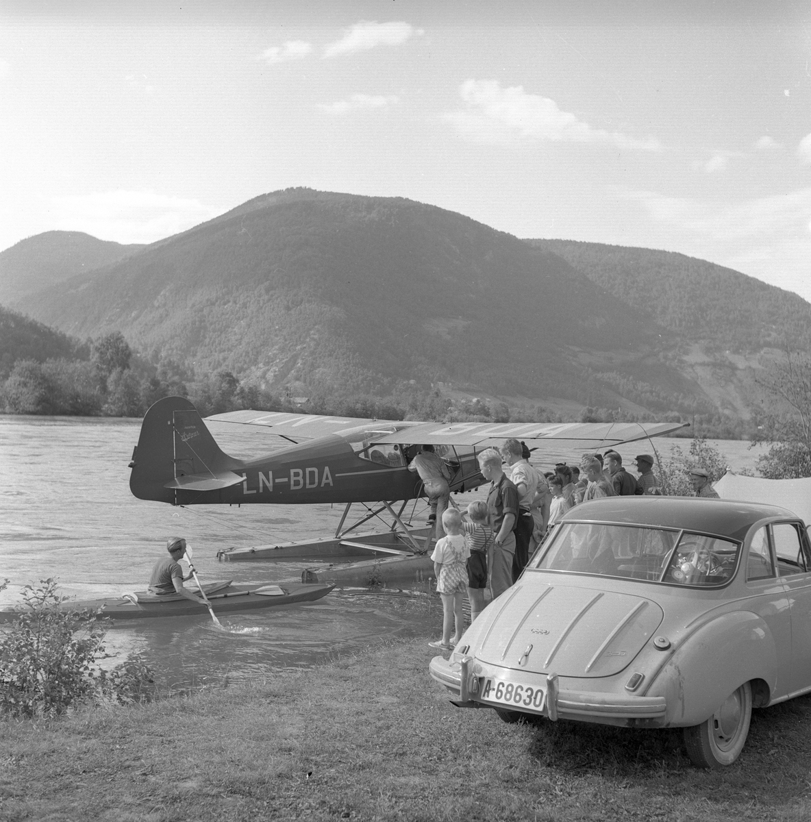 Mann i kajakk og folk som ser på et sjøfly i elva ved Otta. En bil av typen DKW står parkert på elvebredden. Otta, sommeren 1955, campingliv.